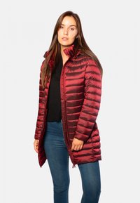 Manteau rembourré rouge avec un design matelassé horizontal, fermeture éclair et capuche. Porté sur un haut noir et un jean bleu, mettant en valeur une texture lisse et brillante.
