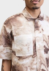 Button-up skjorte i beige og brune nuancer, med tekstureret stof med blomstermønster, to forlommer og krave-design.