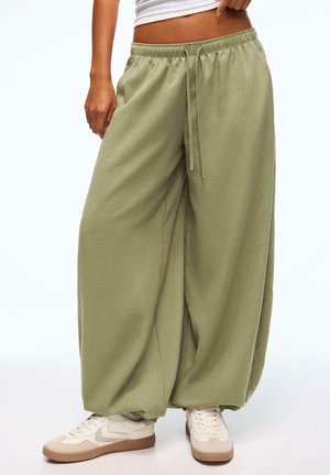 Partie inférieure du corps d'une personne portant un pantalon ample vert clair à cordon et des baskets blanches, debout avec une main reposant près de la taille.