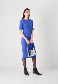Žena stojí v modrých šatech s krátkými rukávy, drží kovovou kabelku a má na sobě černé kotníkové boty, před neutrálním pozadím.