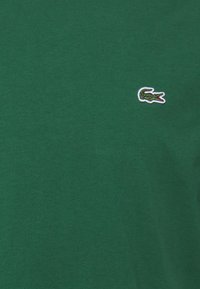 Žalias medvilninis marškinėliai su mažuoju išsiuvinėtu krokodilo logotipu baltoje ir raudonoje spalvose kairiajame krūtinės šone. Lygi audinio tekstūra.