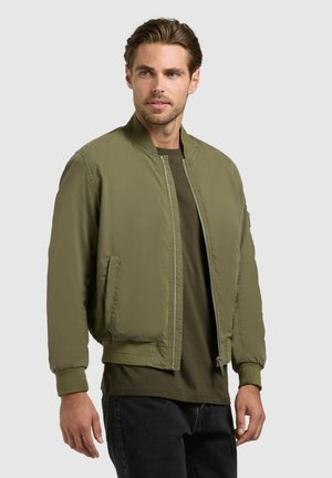 Man wearing olive green bomber jacket over dark green shirt and black pants, standing against plain light gray background.