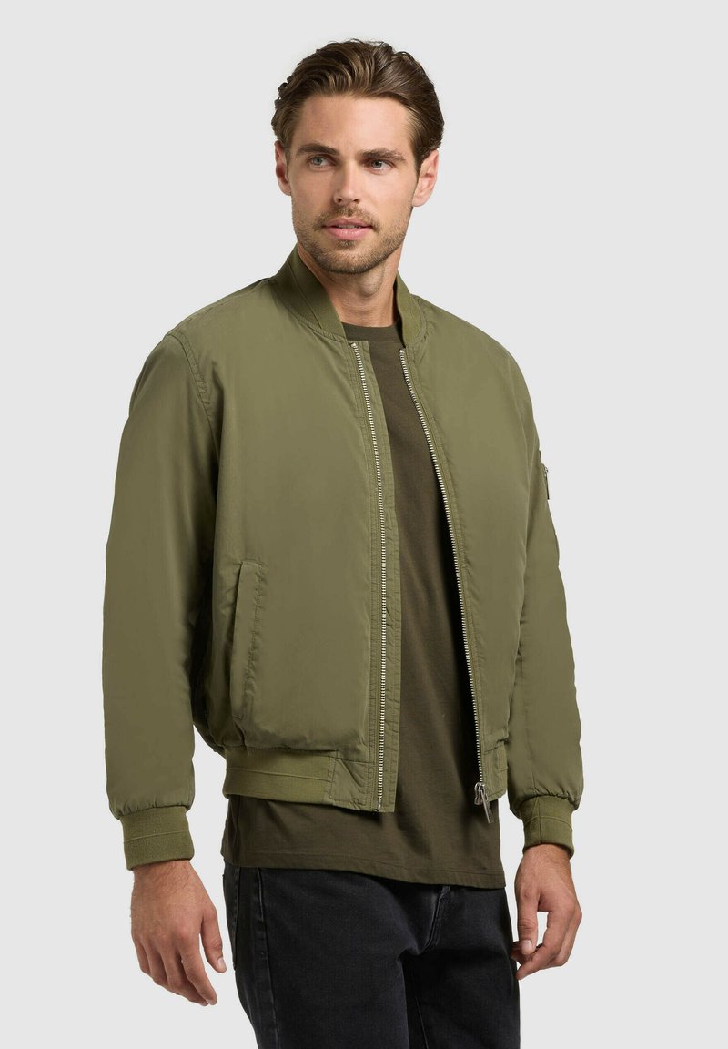 Homme portant une veste bomber vert olive sur une chemise vert foncé et un pantalon noir, debout devant un fond gris clair uni.