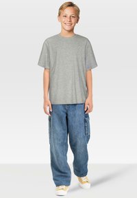 Junge steht lächelnd da, trägt ein graues T-Shirt, lockere blaue Cargo-Jeans und gelbe Sneakers vor einem schlichten hellen Hintergrund.