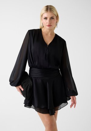 Femme aux cheveux blonds portant une mini robe noire à manches longues, tenant une pochette noire texturée sur un fond gris uni.