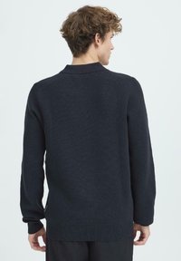 Sötét tengerészkék kötött pulóver hosszú ujjakkal, texturált felülettel és enyhén bordázott szegéllyel. Klasszikus gallérral és oldalsó díszítésekkel.