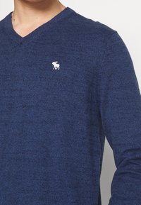 Donkerblauwe V-halstrui gemaakt van een textuur gebreide stof, met een klein wit액el logo op de linkerborst.