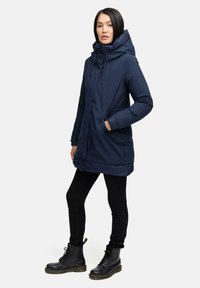 Marineblaue, isolierte Parka mit hohem Kragen, Kapuze mit Kordelzug, seitlichen Taschen und elastischen Akzenten an der Taille. Schwarze Skinny-Jeans und Kampfstiefel.