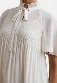 Blouse blanche avec des plis verticaux, un col montant et une fermeture à nœud. Tissu doux et lisse avec des manches courtes et un design fluide et froncé.