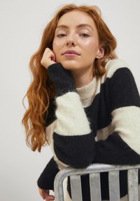 Gestreifter Pullover in Schwarz und Creme mit geripptem Ausschnitt und strukturierter Stoffqualität. Das Model stützt ihr Kinn auf ihre Hand gegen einen Metallstuhl.
