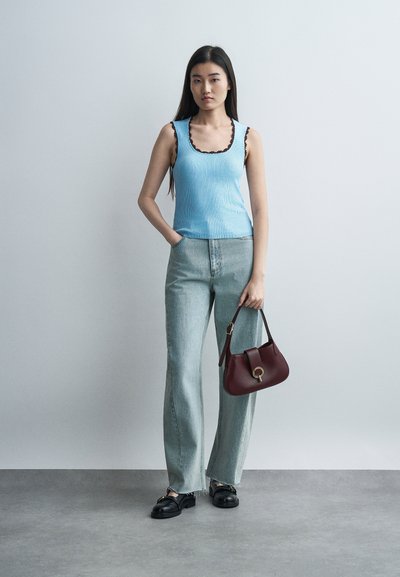 Νεαρή γυναίκα στέκεται μπροστά σε απλό φόντο φορώντας μπλε αμάνικο τοπ, ανοιχτόχρωμα τζιν, μαύρα μοκασίνια και κρατώντας μια μπορντό τσάντα στο δεξί της χέρι.