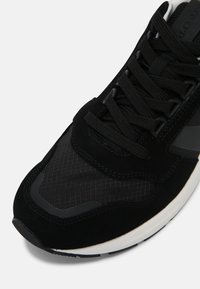 Chaussure de sport noire combinant du daim et du mesh, avec un embout texturé et une semelle extérieure blanche à motif.