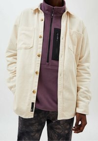 Katoenen overhemd in crème met bruine knopen, gelaagd over een paarse fleece-top met een zwarte ritszak, gedragen met bedrukte donkere broek.