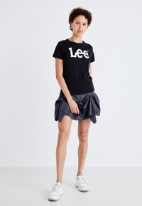 Melnas kokvilnas T-krekls ar baltu "Lee" logo, kombinēts ar pelēku, locītu mini svārku. Modelis valkā baltas masīvas sporta kurpes. Neitrāls fons.