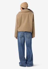 Bēša flīsa jaka ar apkakli, sānu kabatām un brīvu piegriezumu, kopā ar gaiši zilām platām džinsa biksēm un bēšu tamborētu cepuri.