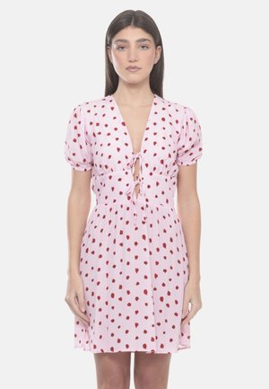 Donna che indossa un vestito rosa chiaro con pois rossi, maniche corte a sbuffo, scollo a V profondo e chiusure con fiocco sul davanti.