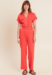 Jumpsuit corail avec des manches courtes retroussées, devant à boutons, deux poches poitrine et taille ceinturée ; conception à jambes larges avec un tissu texturé.