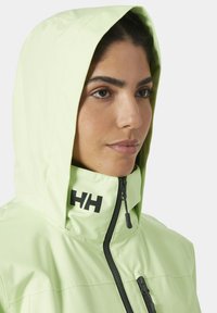 Chaqueta impermeable de color verde claro con capucha, cremallera negra y acento de logo negro en el cuello. Textura suave y diseño ajustado.