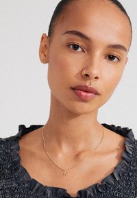 Collana d'oro con un ciondolo a forma di lettera "N", indossata su un top scuro con texture e un colletto a volant. Pelle liscia e trucco naturale.