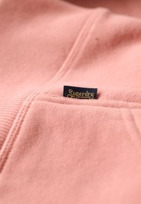 Chaqueta de forro polar rosa con bolsillo lateral, con una etiqueta azul marino con el texto dorado "Superdry". Textura suave con un acabado liso.