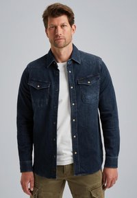 Ciemnoniebieska koszula dżinsowa z zapięciem na guziki z przodu, kieszeniami na piersi i kontrastowymi szwami. Nosi się ją na białym t-shircie i zestawia z spodniami cargo.