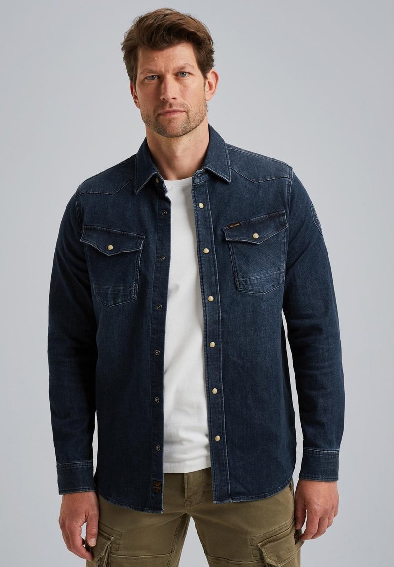 Ciemnoniebieska koszula dżinsowa z zapięciem na guziki z przodu, kieszeniami na piersi i kontrastowymi szwami. Nosi się ją na białym t-shircie i zestawia z spodniami cargo.