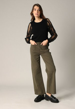Femme se tenant avec les mains dans les poches, portant un haut noir transparent à manches longues, un pantalon large olive et des mocassins noirs épais sur fond gris.