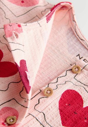 Ružová látka s červenými srdiečkovými vzormi, má textúrovaný povrch, preklopený golier a dva svetlohnedé gombíky blízko detailov šitia.