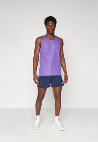 Lila Sporttanktop mit einem strukturierten Muster, kombiniert mit marineblauen Shorts und hellgrauen Laufschuhen mit grünen Akzenten.