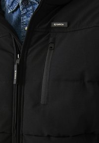 Chaqueta de plumas negra con un bolsillo con cremallera, con una textura suave y una etiqueta con el logo. Usada sobre una camisa de mezclilla azul.