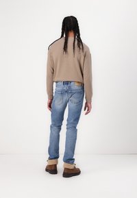Sudadera beige, vaqueros de mezclilla azules, botas marrones con forro de piel; persona vista desde atrás, con trenzas, contra un fondo liso.