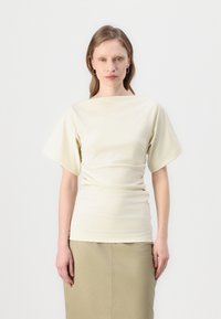 Blouse crème avec des manches courtes et larges et un col bateau. Le tissu a une texture lisse, drapé à la taille, associé à une jupe beige.