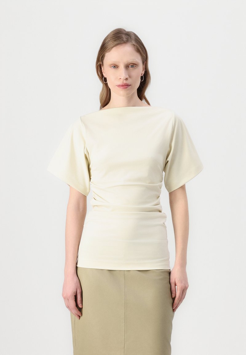 Blouse crème avec des manches courtes et larges et un col bateau. Le tissu a une texture lisse, drapé à la taille, associé à une jupe beige.