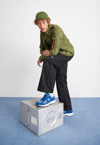 Green utility jacket with zip pocket, black wide-leg pants, and blue New Balance sneakers on a metallic crate against a plain background.