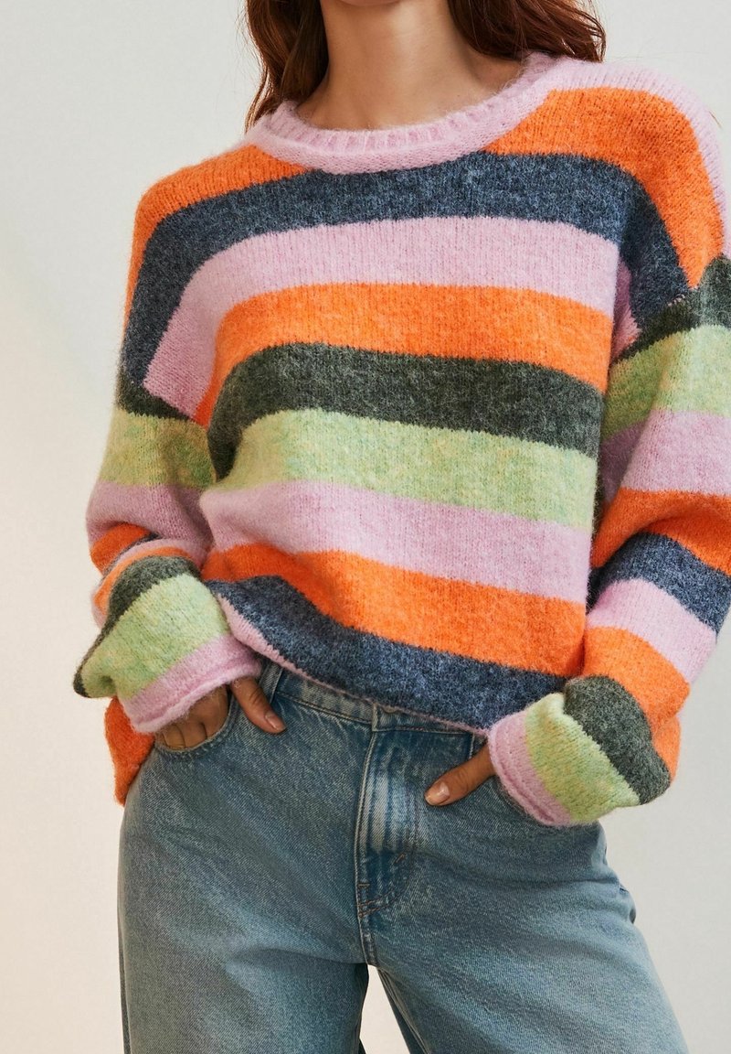 Femme portant un pull douillet à larges rayures horizontales orange, rose, gris foncé et vert, assorti à un jean en denim bleu.