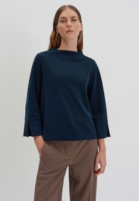 Navy blue, lightweight top with a high neckline, three-quarter sleeves, and side slits. Smooth texture with a relaxed fit, paired with beige trousers.