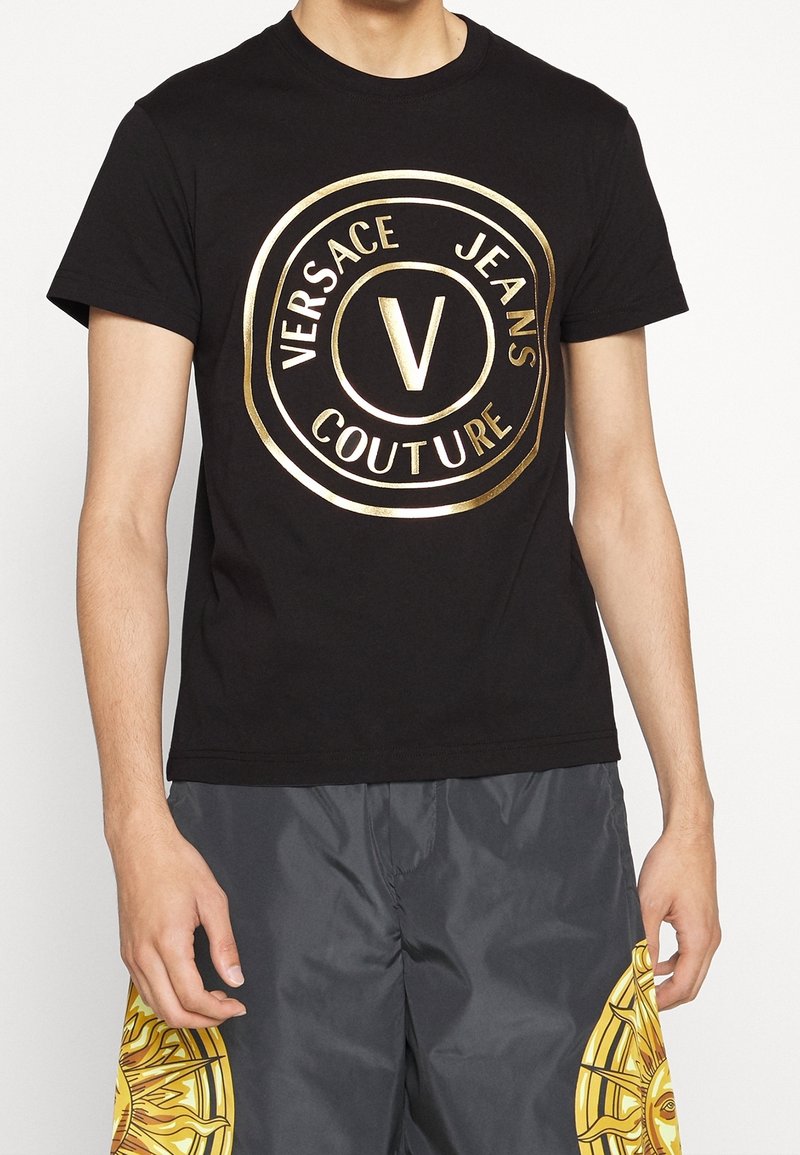 Schwarzes Baumwoll-T-Shirt mit einem auffälligen, goldenen runden Logo, das "VERSACE JEANS COUTURE" und ein zentrales "V"-Design zeigt. Kurze Ärmel, Rundhalsausschnitt.