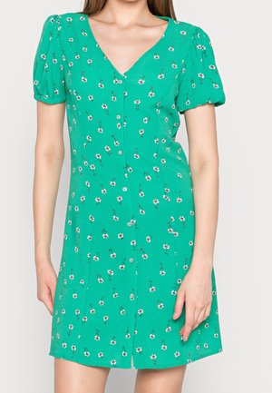 Grøn kjole med knapper, lille hvidt blomsterprint, puffede korte ærmer og V-hals båret af en stående person.