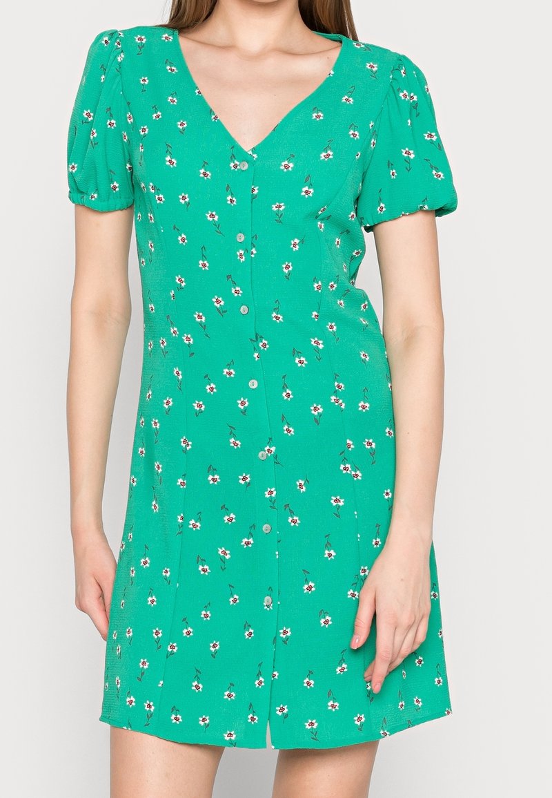 Robe chemise verte à petites fleurs blanches, manches courtes bouffantes et décolleté en V portée par une personne debout.
