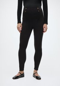 Γυναίκα που φοράει μαύρο ψηλόμεσο στενό παντελόνι με χρυσά κουμπιά και μαύρες γυαλιστερές μπαρετάτες Mary Jane, στέκεται μπροστά σε λευκό φόντο.