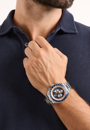 Férfi, aki a gallérját igazítja, texturált tengerészkék pólóinget és ezüst kronográf órát visel kék és narancssárga részletekkel.