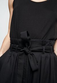 Haut sans manches noir avec une large ceinture, doté d'une ceinture nouée. Fabriqué en tissu lisse, avec une silhouette simple et épurée.