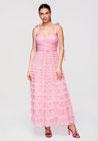 Femme portant une robe en tulle rose à volants sans manches avec des liens rubans aux épaules et des sandales à talons hauts violettes à brides, debout devant un fond uni.