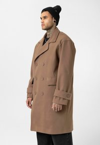 Man met tattoos op nek en hand draagt een bruin mantelpak met dubbele rij knopen, een zwarte muts en een zwarte broek, staande tegen een effen achtergrond.