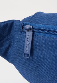Bolsa de tecido azul com uma superfície texturizada e um fecho de correr, apresentando uma pega rectangular da Reebok. A vista em close destaca os detalhes e a costura.