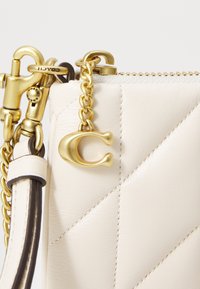 Roh krémové kožené prošívané tašky se zlatým řetízkem, zipem a zlatým přívěskem "C" připojeným poblíž koženého popruhu.