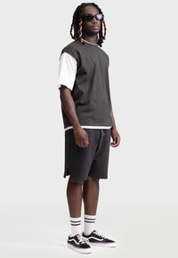 Tricou stratificat gri și alb cu mâneci scurte, asortat cu pantaloni scurți negri și încălțăminte Vans negre. Modelul poartă ochelari de soare și șosete cu dungi.