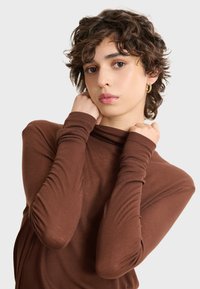 Smeđa turtleneck majica izrađena od mekog rebrastog materijala, s dugim rukavima i naborima na vratu. Glatka tekstura, ležeran kroj.