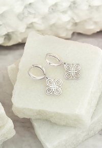 Silberne Creolen mit schmetterlingsförmigen Anhängern, die ein Durchbruchsdesign und eine polierte Oberfläche aufweisen, auf einem hellen Stein platziert.