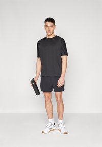 Svart idrettskjorte med korte ermer og en avslappet passform, kombinert med marineblå shorts. Holder en svart vannflaske, iført hvite joggesko.